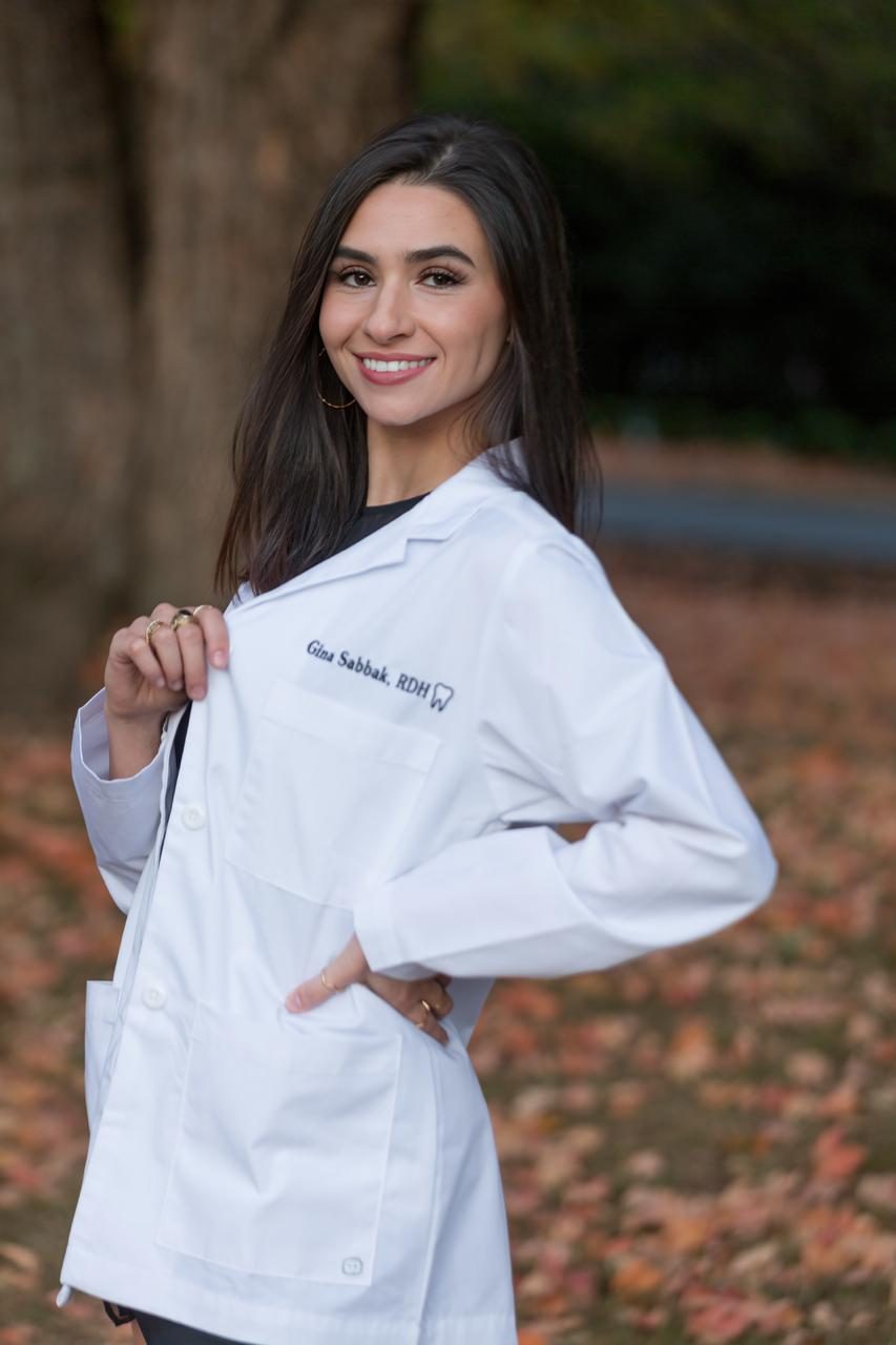 Gina Sabbak, RDH — Myofunctional Therapist, in a white coat with embroidery reading 'Gina Sabbak RDH'.